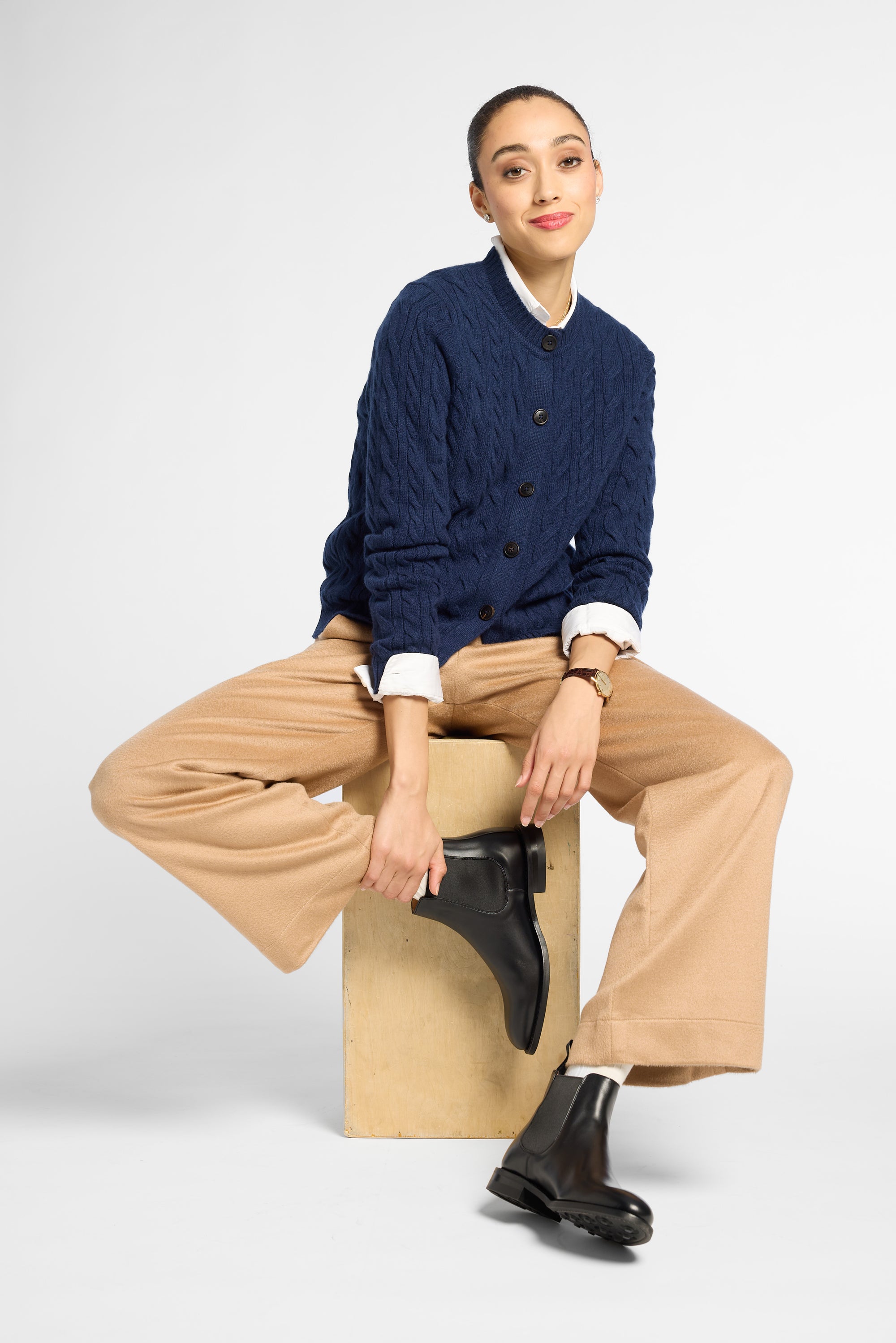 A model sits on a light wood block wearing a navy blue cable-knit cardigan over a white collared shirt, beige wide-leg trousers, and black Chelsea boots, against a light gray studio background.