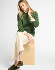 A woman sits on a light wood block wearing a green knit sweater over a white collared shirt, cream wide-leg trousers, and dark olive loafers, photographed against a clean light gray studio background.