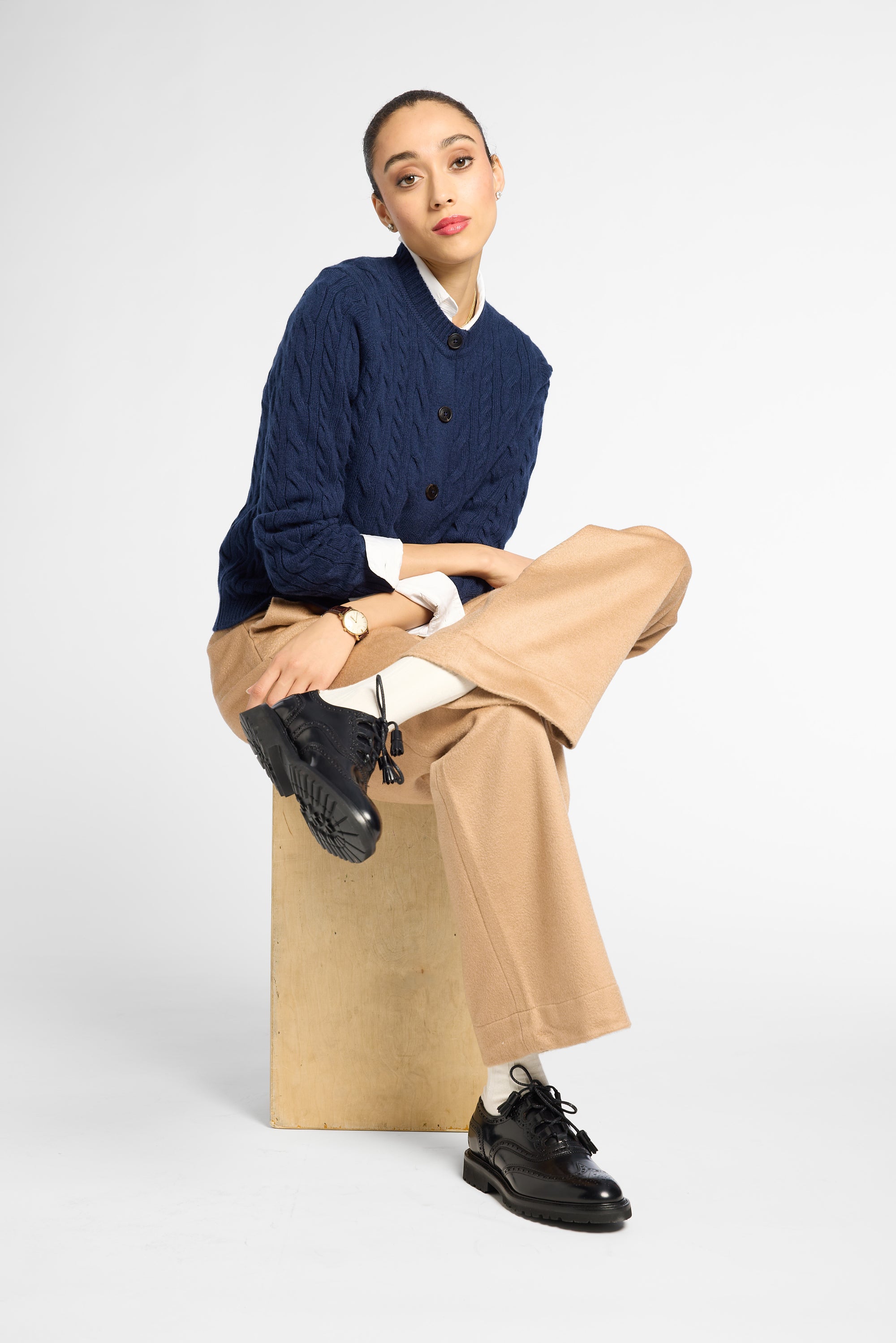A model sits on a light brown cardboard box wearing a navy blue cable-knit cardigan over a white collared shirt, paired with beige wide-leg trousers and black leather shoes, against a plain white studio background.