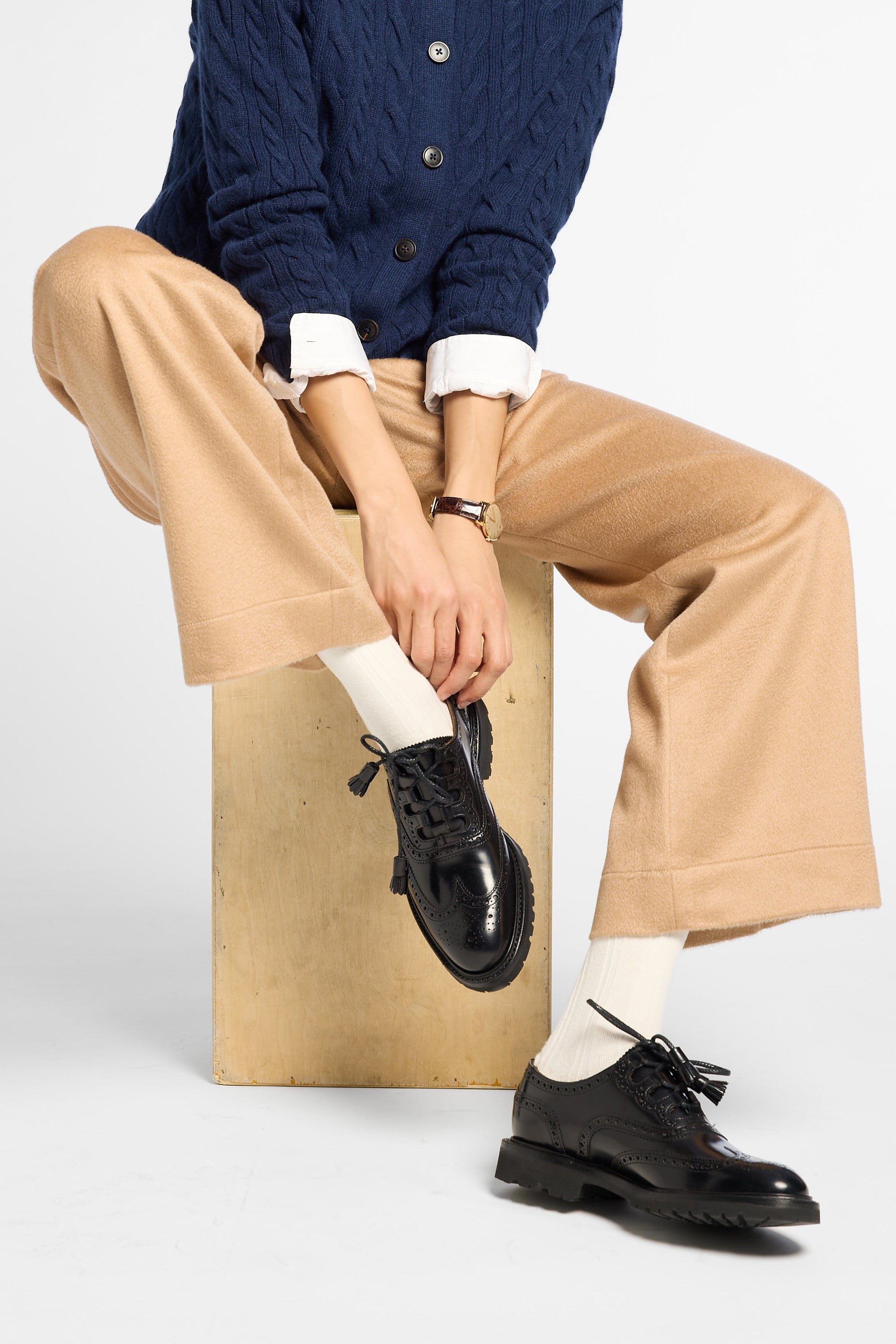 A model sits on a wooden block wearing a navy blue cable-knit cardigan, beige wide-leg trousers, white socks, and black leather oxford shoes against a light gray studio background.