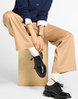 A model sits on a wooden block wearing a navy blue cable-knit cardigan, beige wide-leg trousers, white socks, and black leather oxford shoes against a light gray studio background.