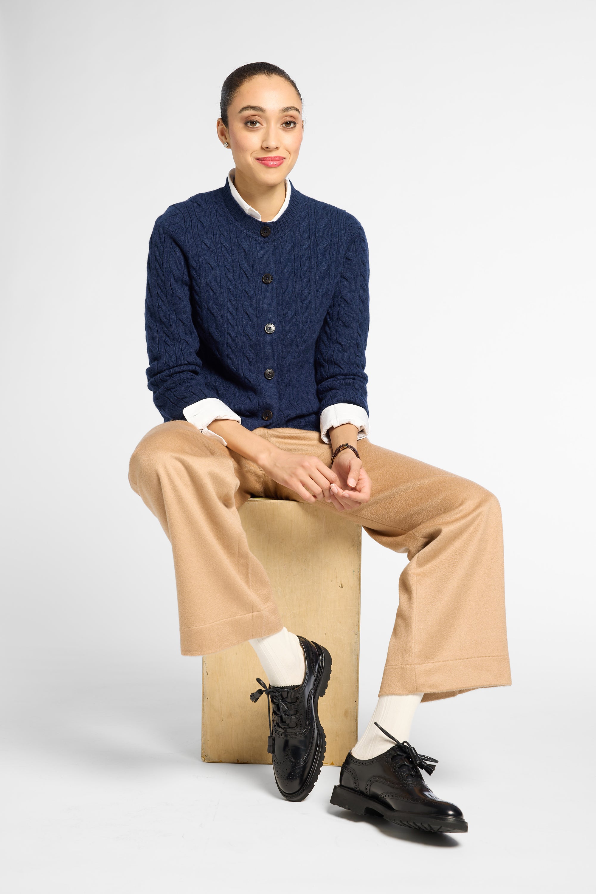 A model sits on a cardboard box wearing a navy blue cable-knit cardigan over a white collared shirt, beige wide-leg trousers, white socks, and black leather shoes against a light gray studio background.