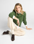A woman sits on a light gray studio floor wearing a green knit sweater over a white collared shirt, cream wide-leg trousers, and dark brown leather loafers.