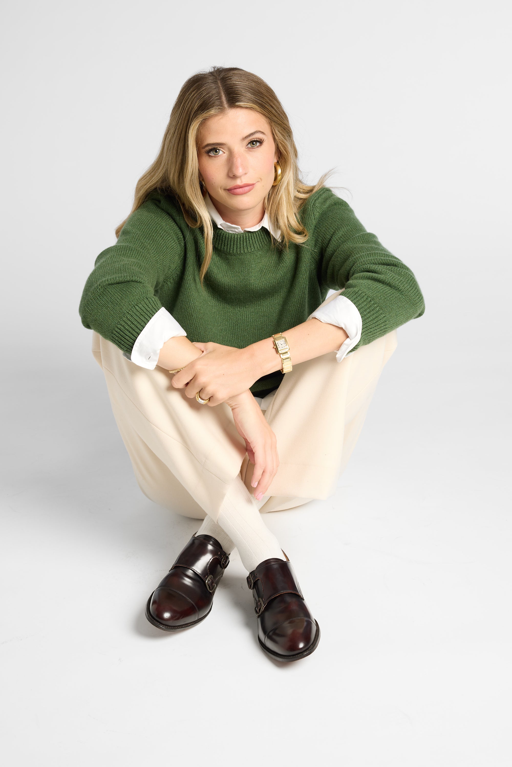 A woman crouching in a studio setting, wearing a green knit sweater over a white collared shirt, cream trousers, and dark brown leather loafers.