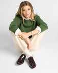 A woman crouching in a studio setting, wearing a green knit sweater over a white collared shirt, cream trousers, and dark brown leather loafers.