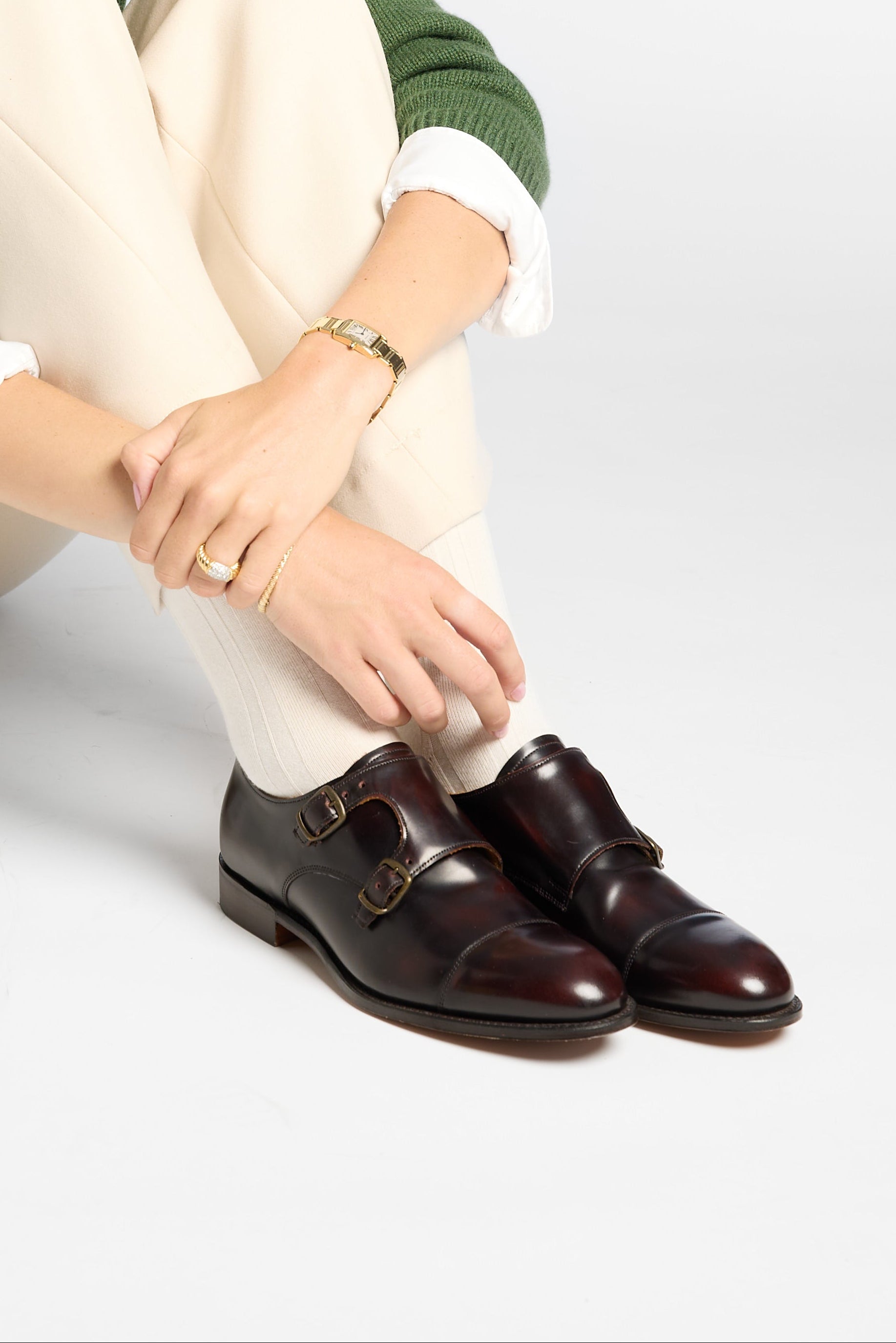 A woman sits cross-legged on a light gray studio floor wearing a green knit sweater over a white collared shirt, cream pants, and dark brown leather loafers.
