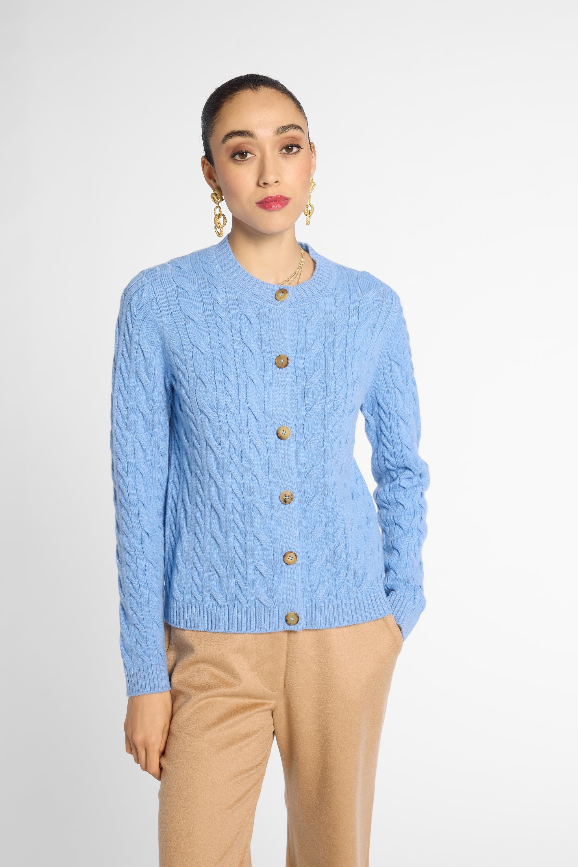 A woman modeling a light blue cable-knit cardigan sweater with brown buttons, paired with beige trousers, against a plain light gray studio background.