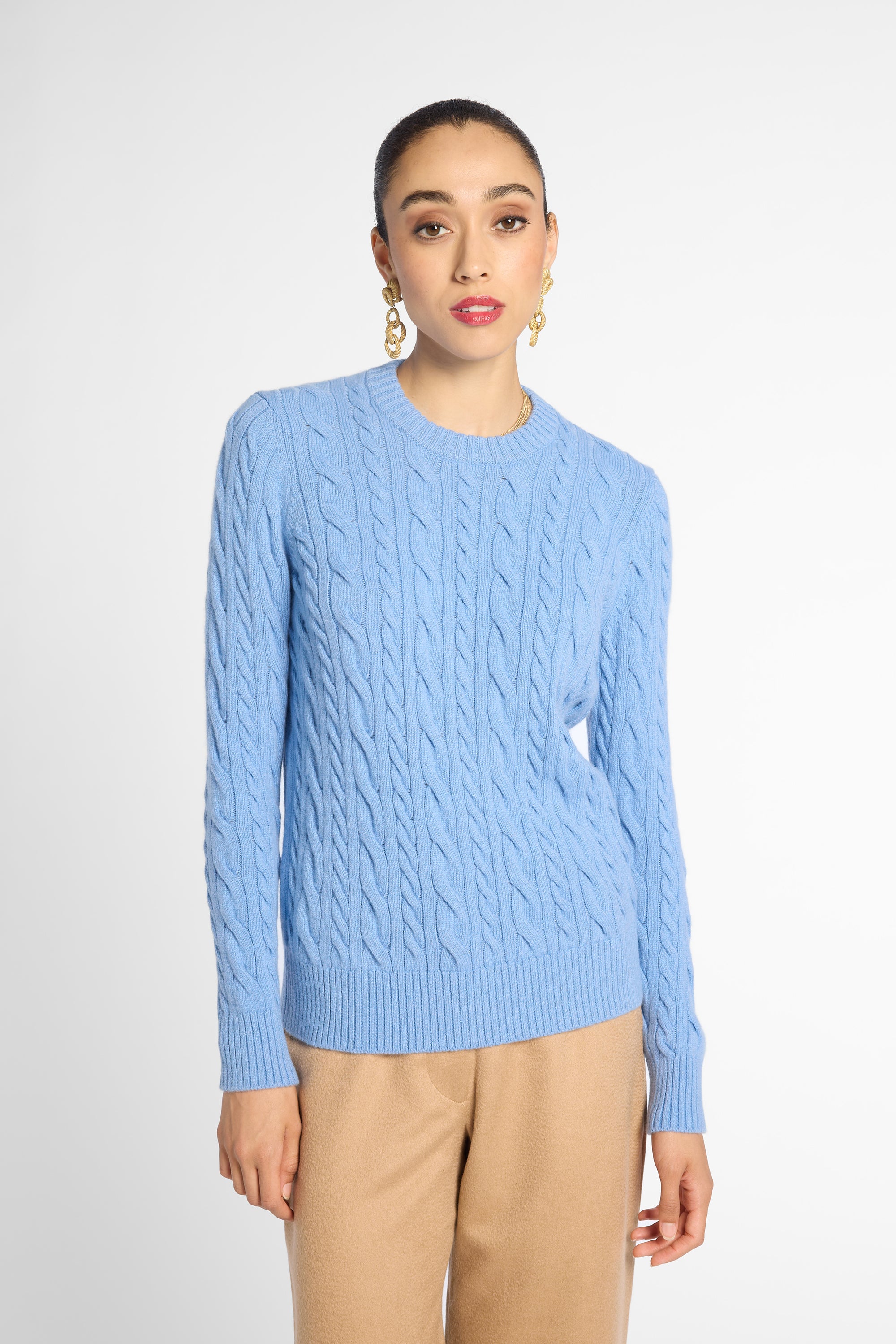 A woman modeling a light blue cable-knit crewneck sweater with beige trousers, standing against a plain light gray studio background.
