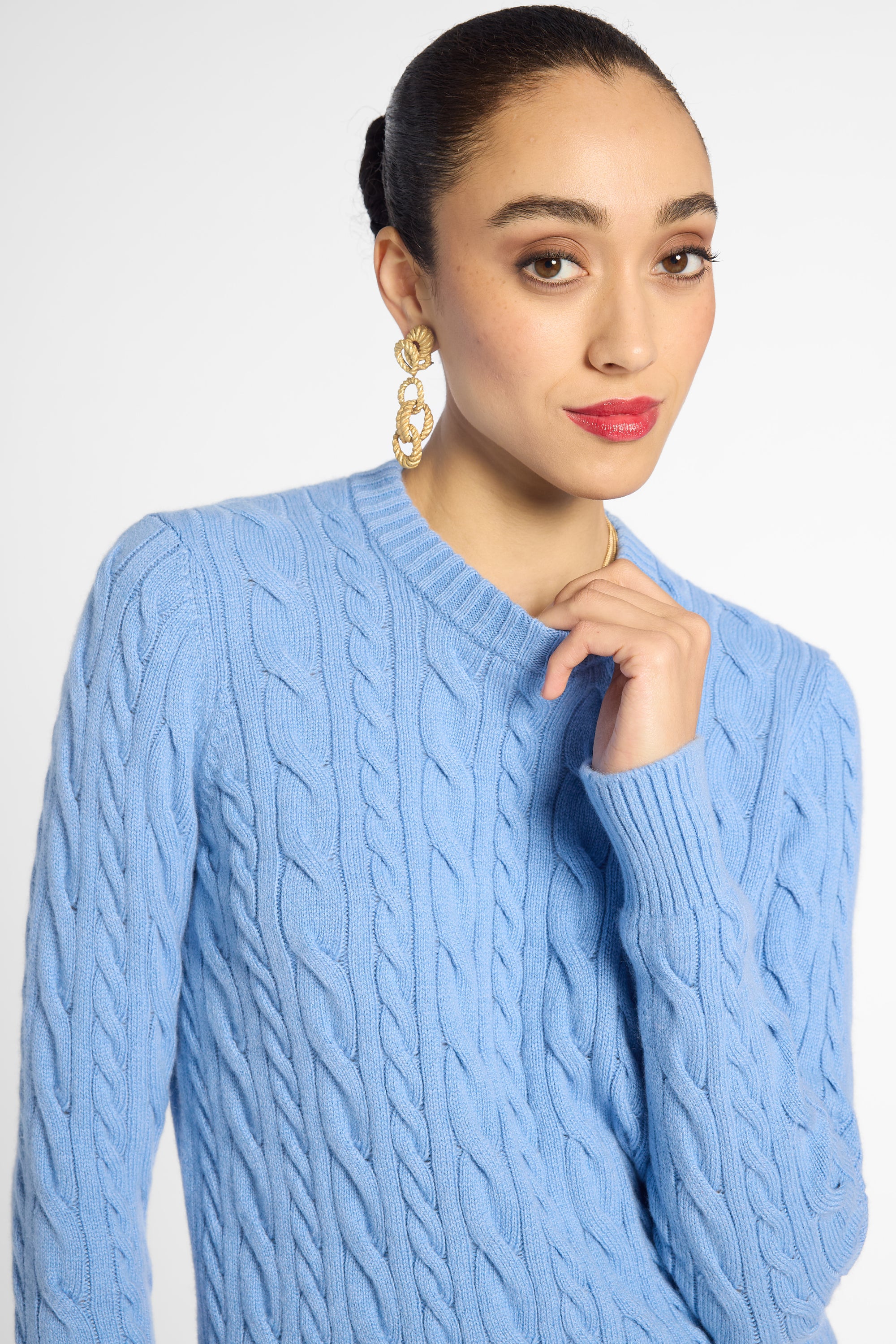 A woman wearing a light blue cable-knit sweater, styled with gold chain earrings, against a plain light gray studio background.