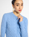 A woman wearing a light blue cable-knit sweater, styled with gold chain earrings, against a plain light gray studio background.