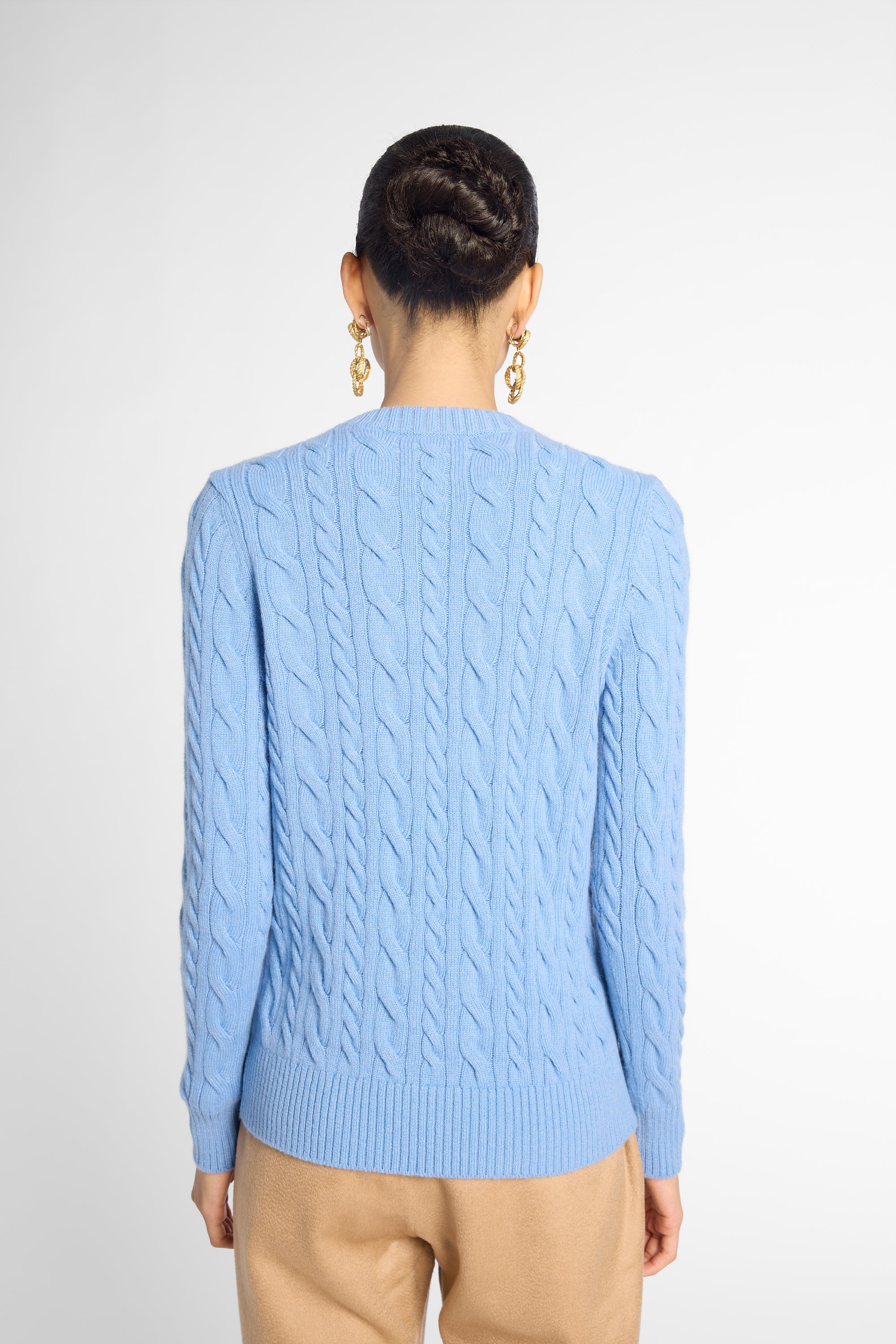 A woman viewed from the back wearing a light blue cable-knit sweater and beige trousers, standing against a plain light gray studio background.