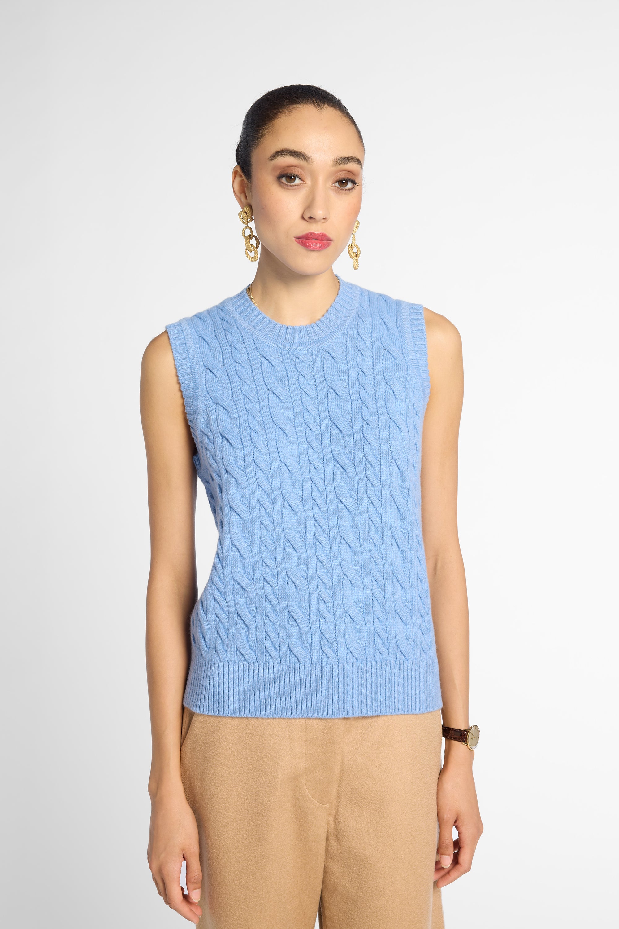 A woman modeling a sleeveless light blue cable-knit sweater vest with beige trousers, gold hoop earrings, and a watch, set against a plain light gray studio background.