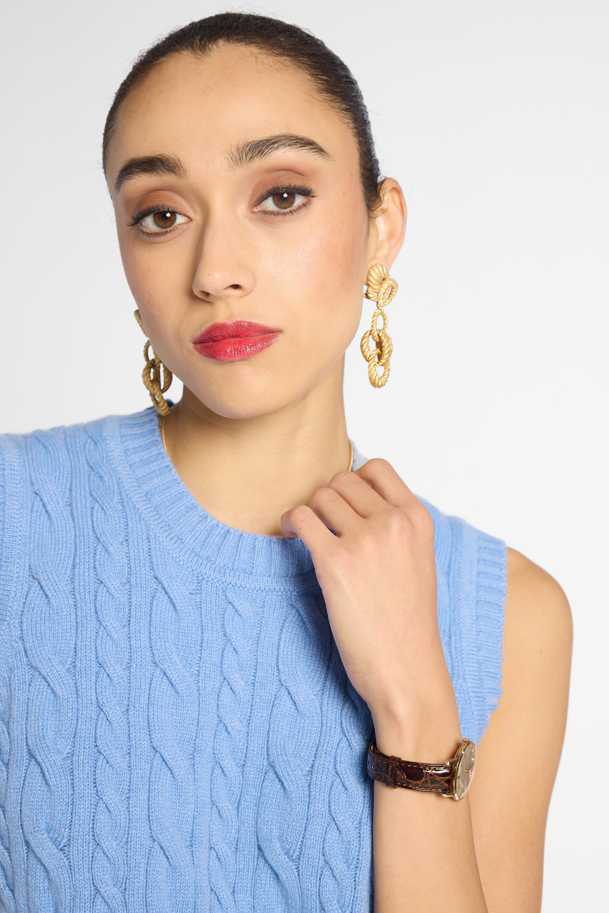 A close-up of a woman wearing gold drop earrings with textured, organic loops, paired with a light blue cable-knit sweater vest and a brown leather watch.