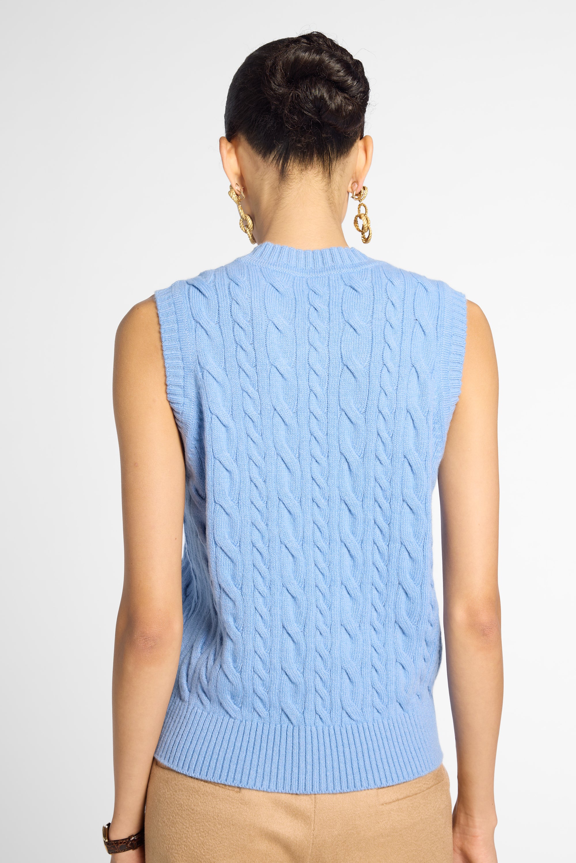 A woman shown from the back wearing a light blue sleeveless cable-knit sweater vest, paired with beige pants, against a plain light gray studio background.