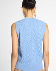 A woman shown from the back wearing a light blue sleeveless cable-knit sweater vest, paired with beige pants, against a plain light gray studio background.