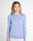 A woman modeling a light blue long-sleeve crewneck sweater, paired with beige pants, against a plain light gray studio background.