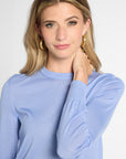 A woman with blonde hair wearing a light blue long-sleeve knit top and gold hoop earrings, posing against a light gray studio background.