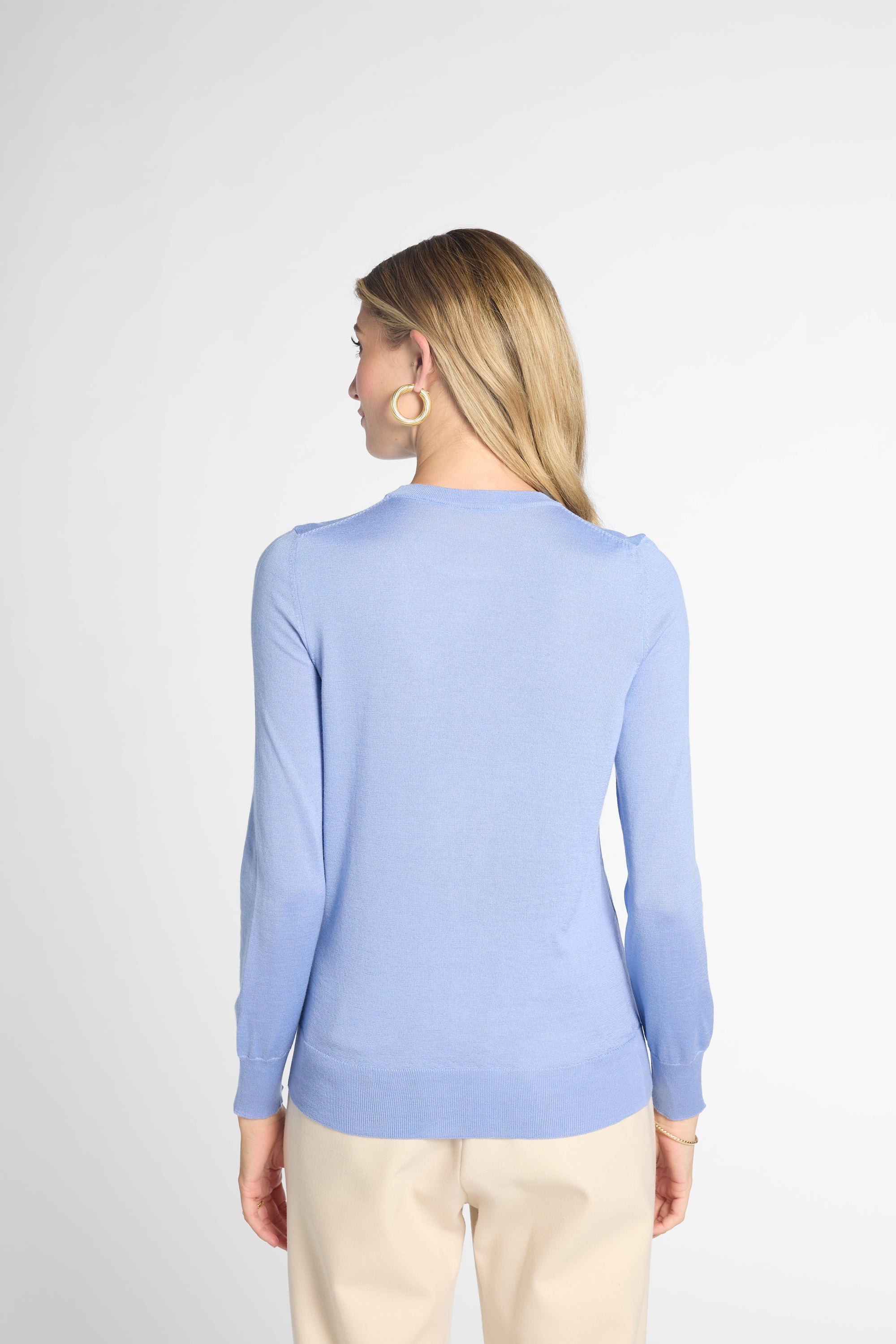 A woman with blonde hair is shown from the back wearing a light blue long-sleeve knit sweater and beige trousers against a light gray studio background.