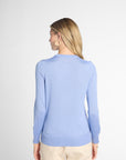 A woman with blonde hair is shown from the back wearing a light blue long-sleeve knit sweater and beige trousers against a light gray studio background.