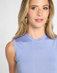A woman with blonde hair wearing a light  blue ribbed sleeveless top and gold hoop earrings, shown in a close-up shot against a light gray background.