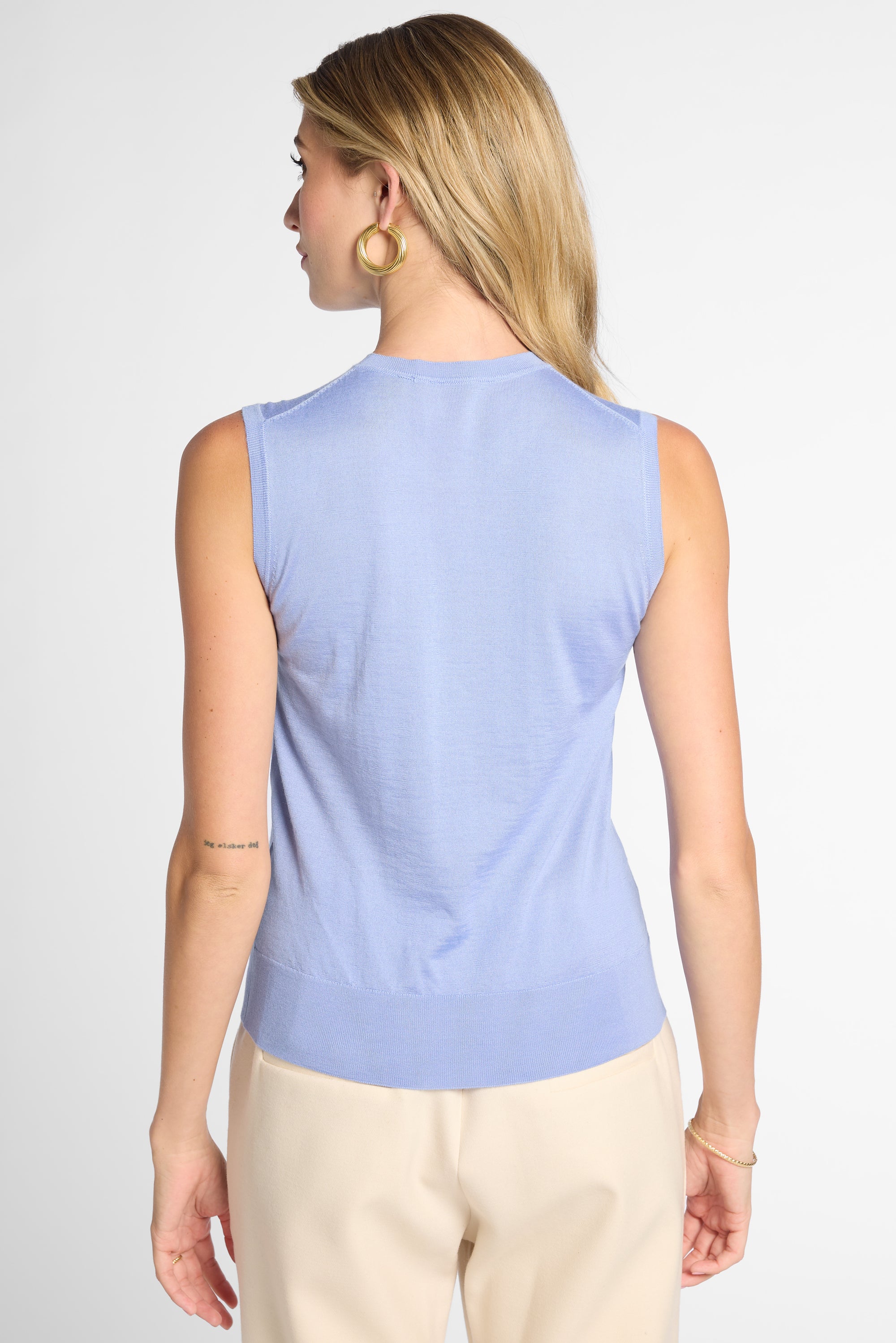 A woman shown from the back wearing a light blue sleeveless knit top and beige pants against a plain light gray studio background.