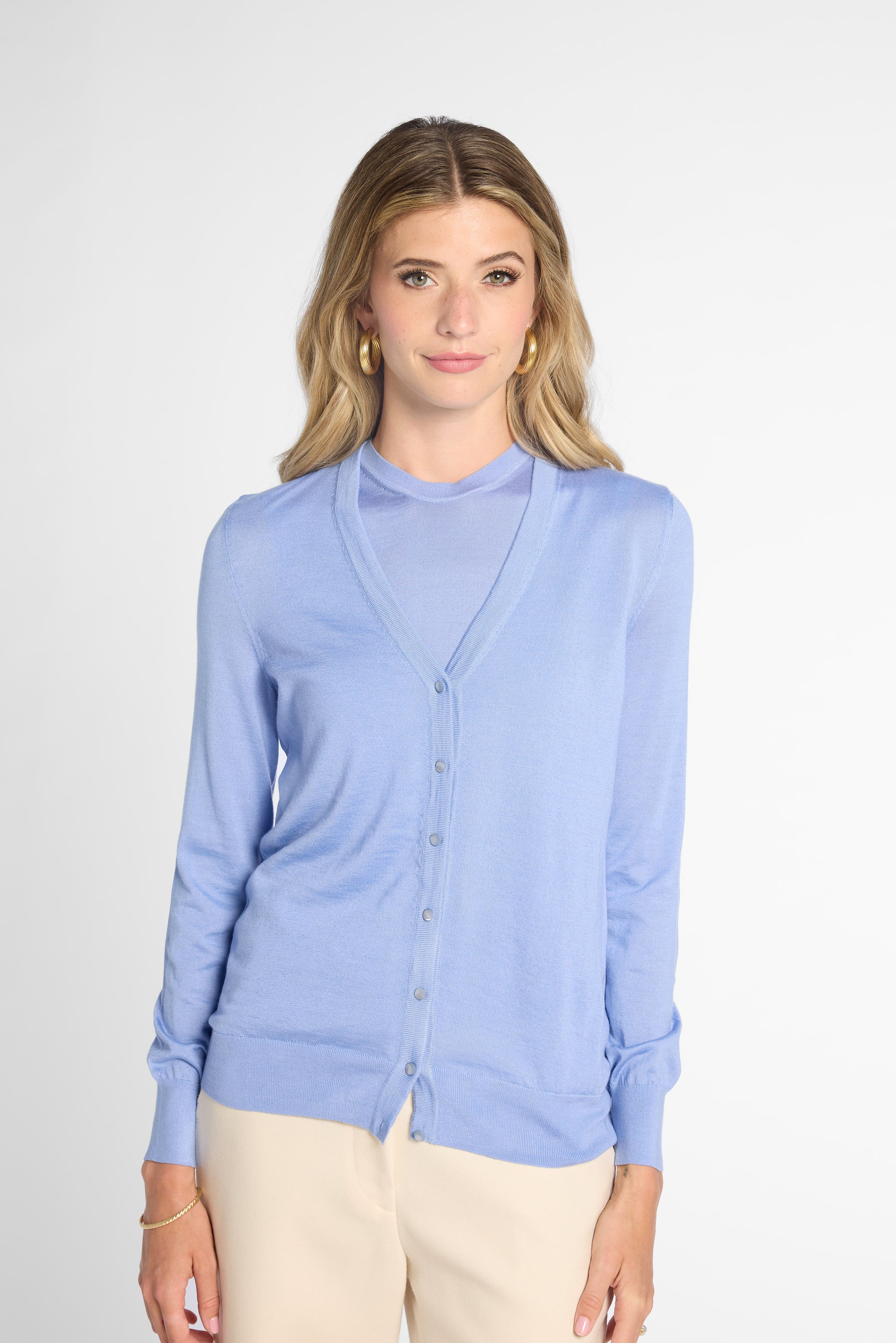 A woman modeling a light blue cardigan sweater and button front, paired with beige pants, against a plain light gray studio background.