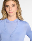 A woman with blonde hair wearing a light blue long-sleeve button-up cardigan over a matching top, paired with gold hoop earrings, against a plain light gray background.