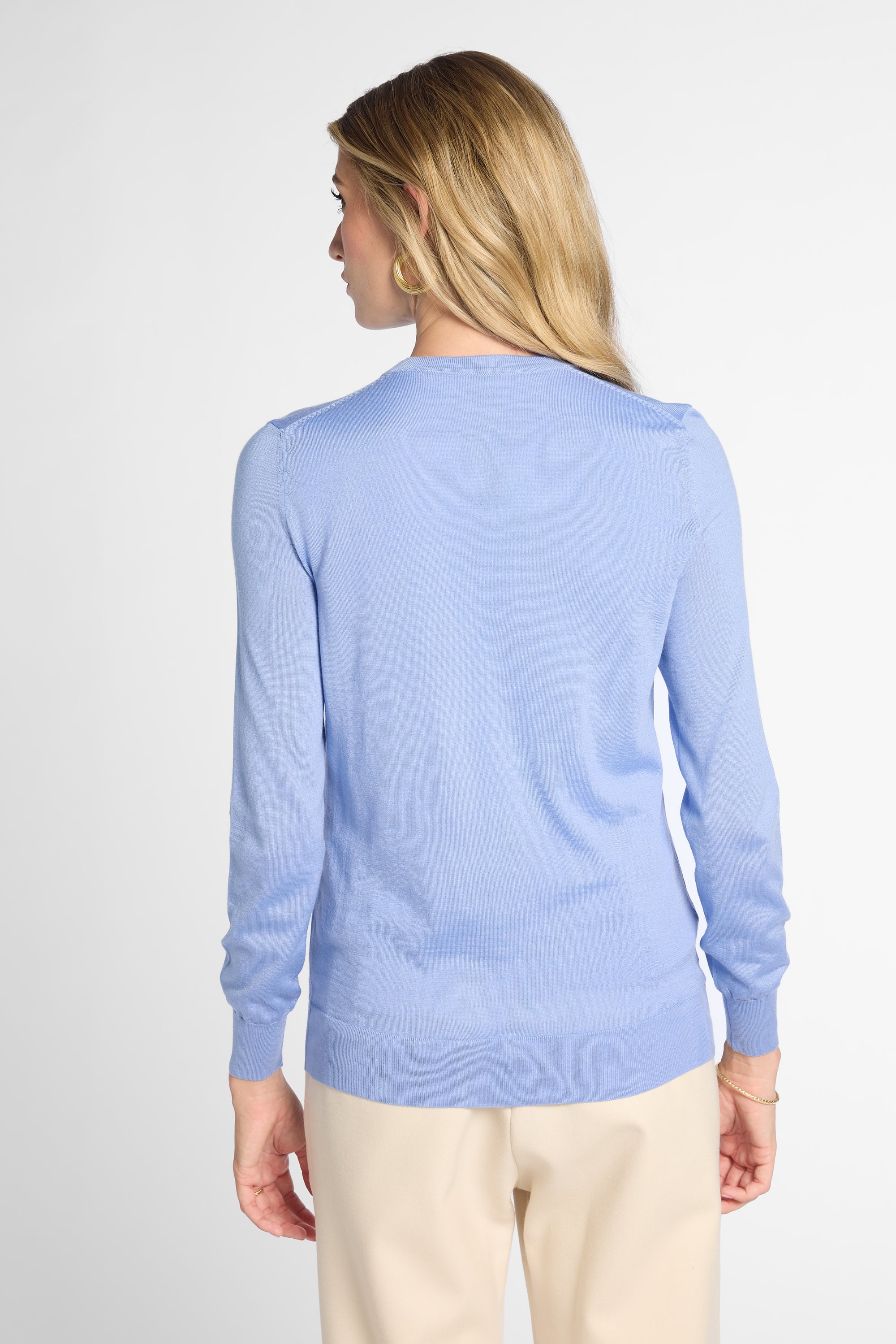 A woman shown from the back wearing a light blue long-sleeve knit sweater and beige pants against a plain light gray studio background.