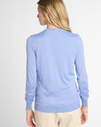A woman shown from the back wearing a light blue long-sleeve knit sweater and beige pants against a plain light gray studio background.