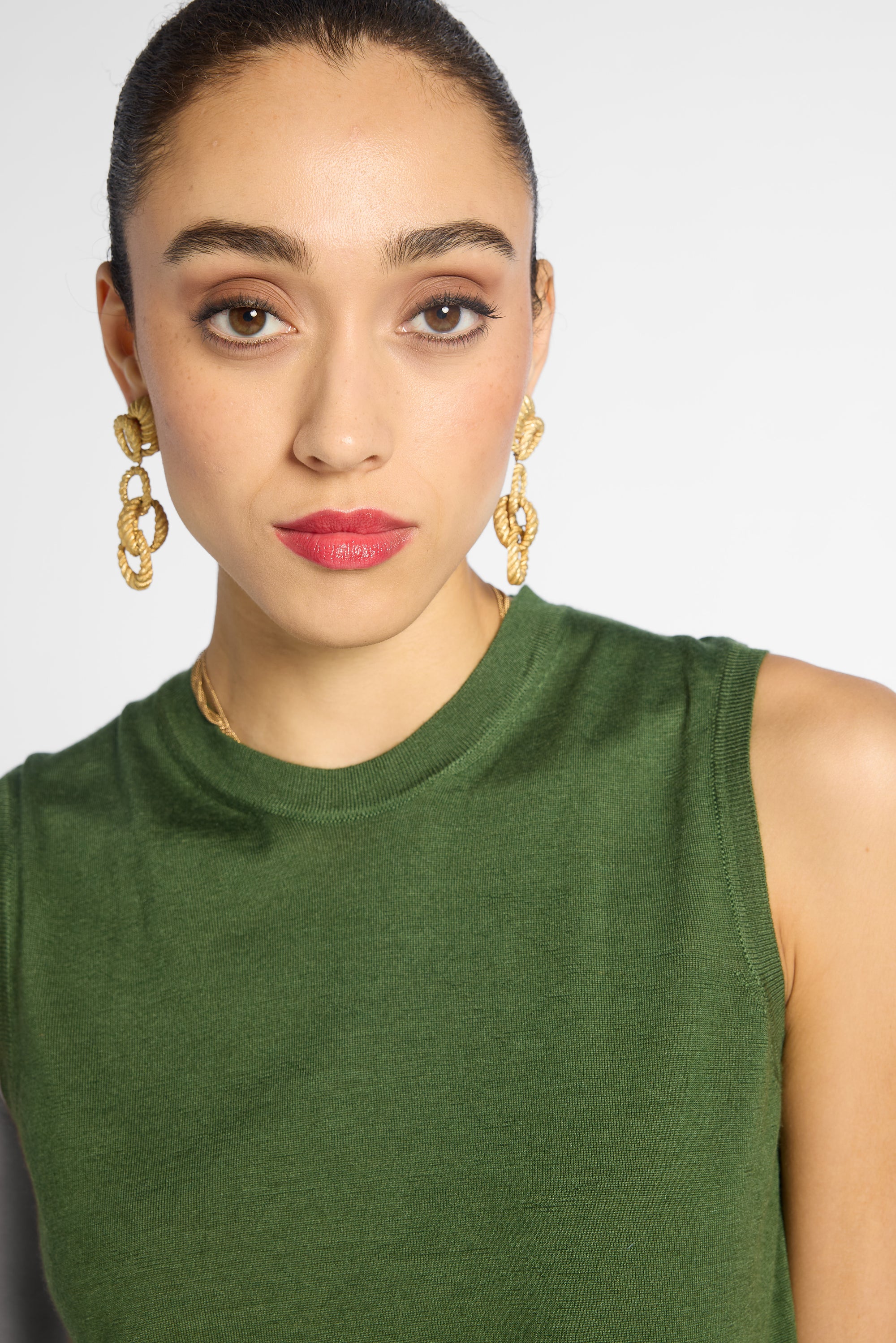 A close-up of a model wearing a sleeveless olive green top and gold chain-link drop earrings against a light gray background.