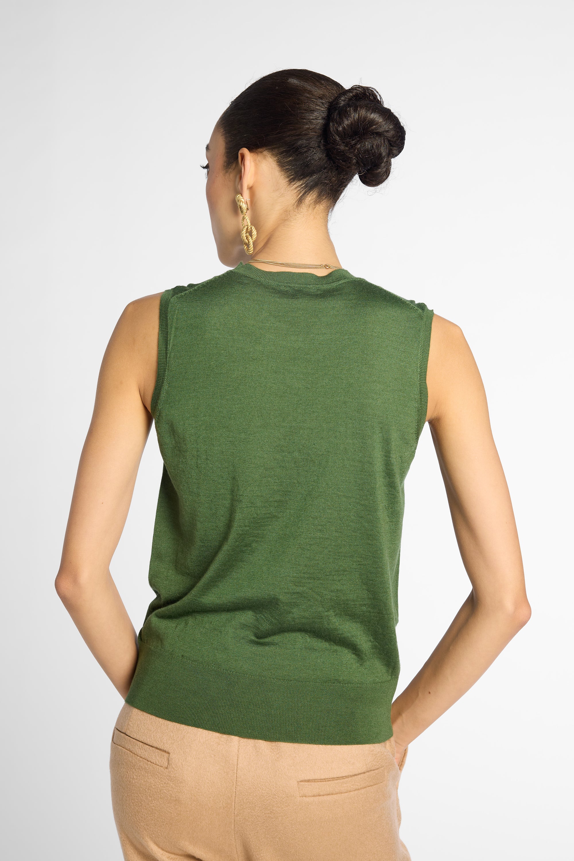 A woman shown from the back wearing a sleeveless green knit top and beige pants against a light gray studio background.