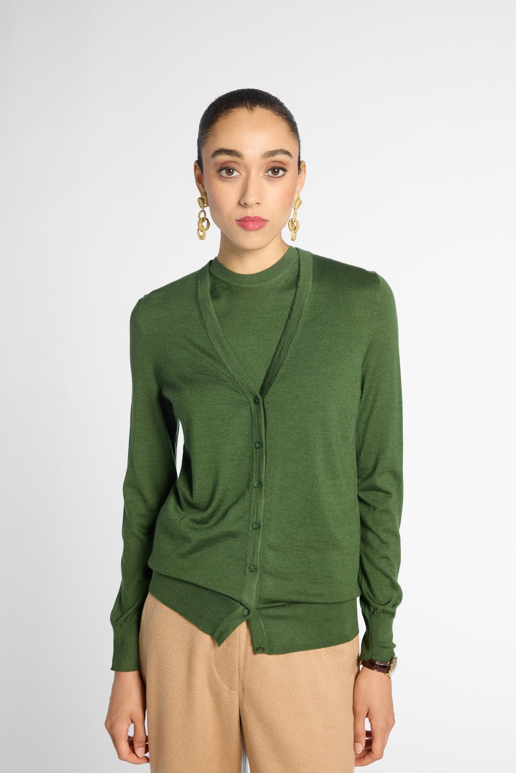 A woman modeling a green long-sleeve button-front cardigan over a matching green top, paired with beige trousers, against a light gray studio background.