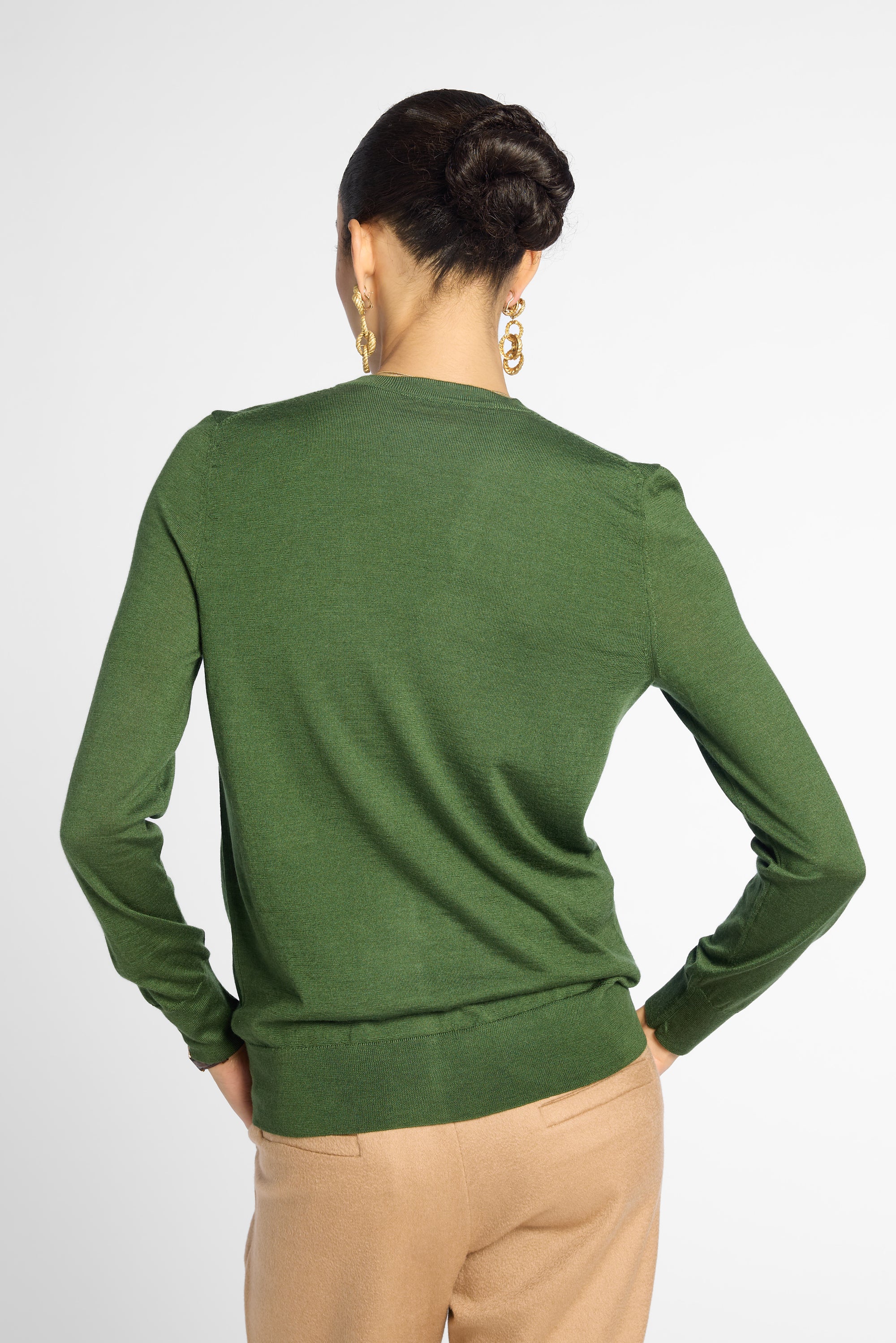 A woman shown from the back wearing a green long-sleeve knit sweater and beige trousers against a light gray studio background.