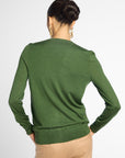 A woman shown from the back wearing a green long-sleeve knit sweater and beige trousers against a light gray studio background.