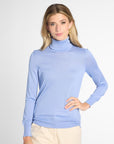 A woman modeling a light blue long-sleeve turtleneck sweater, paired with beige pants, against a plain light gray studio background.