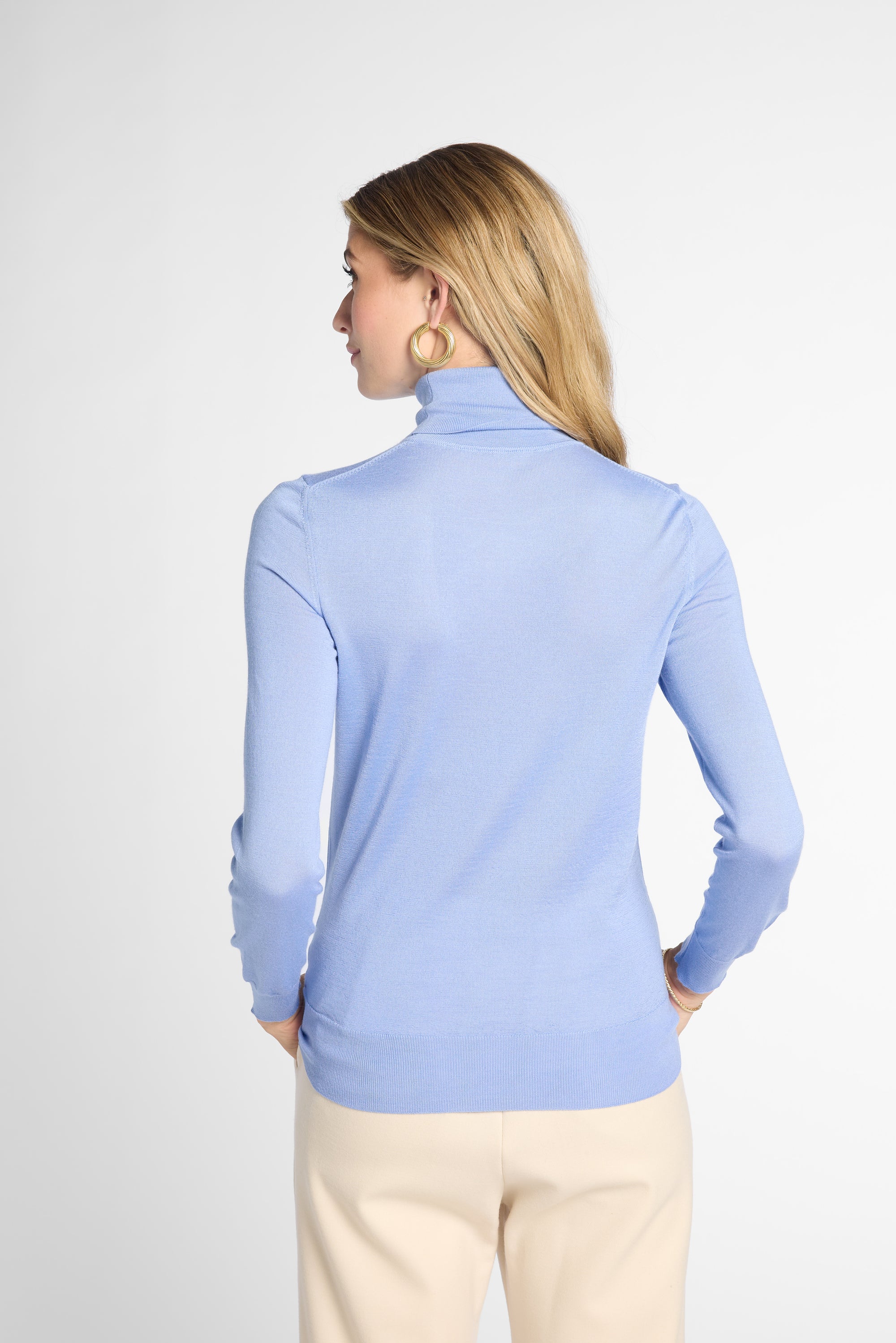 A woman shown from the back wearing a light blue turtleneck sweater and cream pants against a plain light gray studio background.