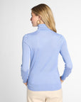 A woman shown from the back wearing a light blue turtleneck sweater and cream pants against a plain light gray studio background.