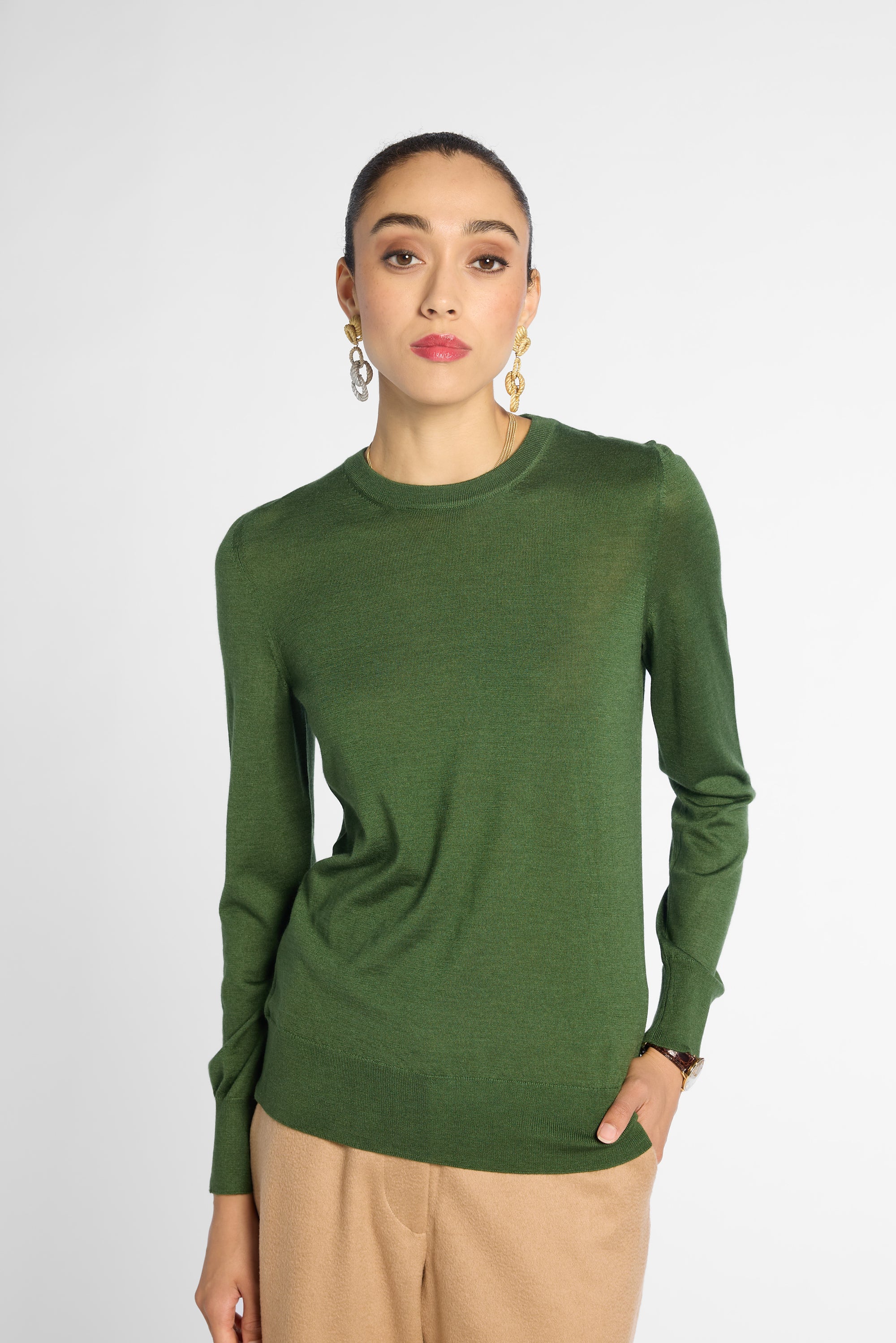 A woman wearing a fitted long-sleeve crewneck sweater in olive green, paired with beige trousers, against a plain light gray studio background.