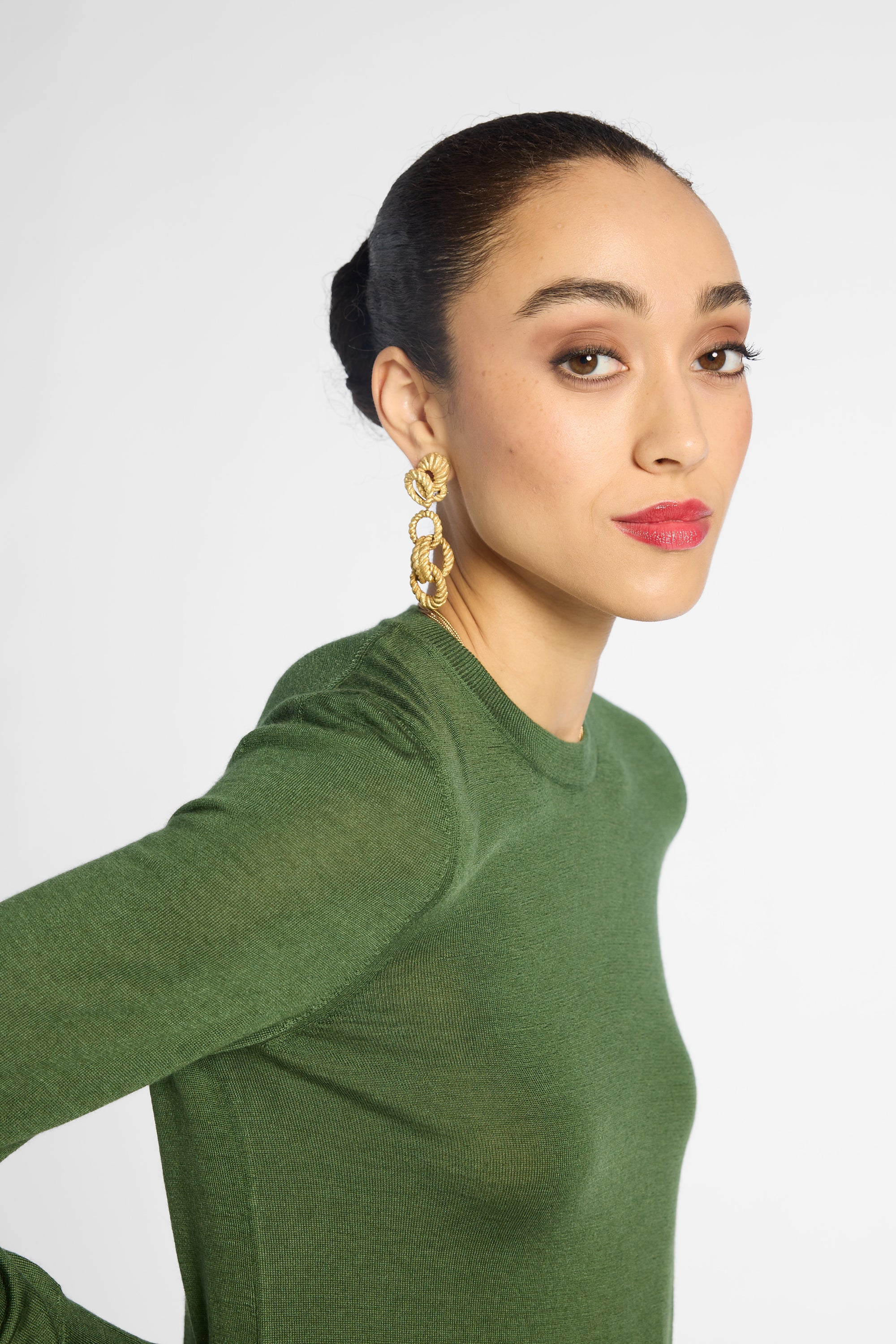 A woman wearing a green long-sleeve knit top and large gold statement earrings, photographed against a light gray studio background.