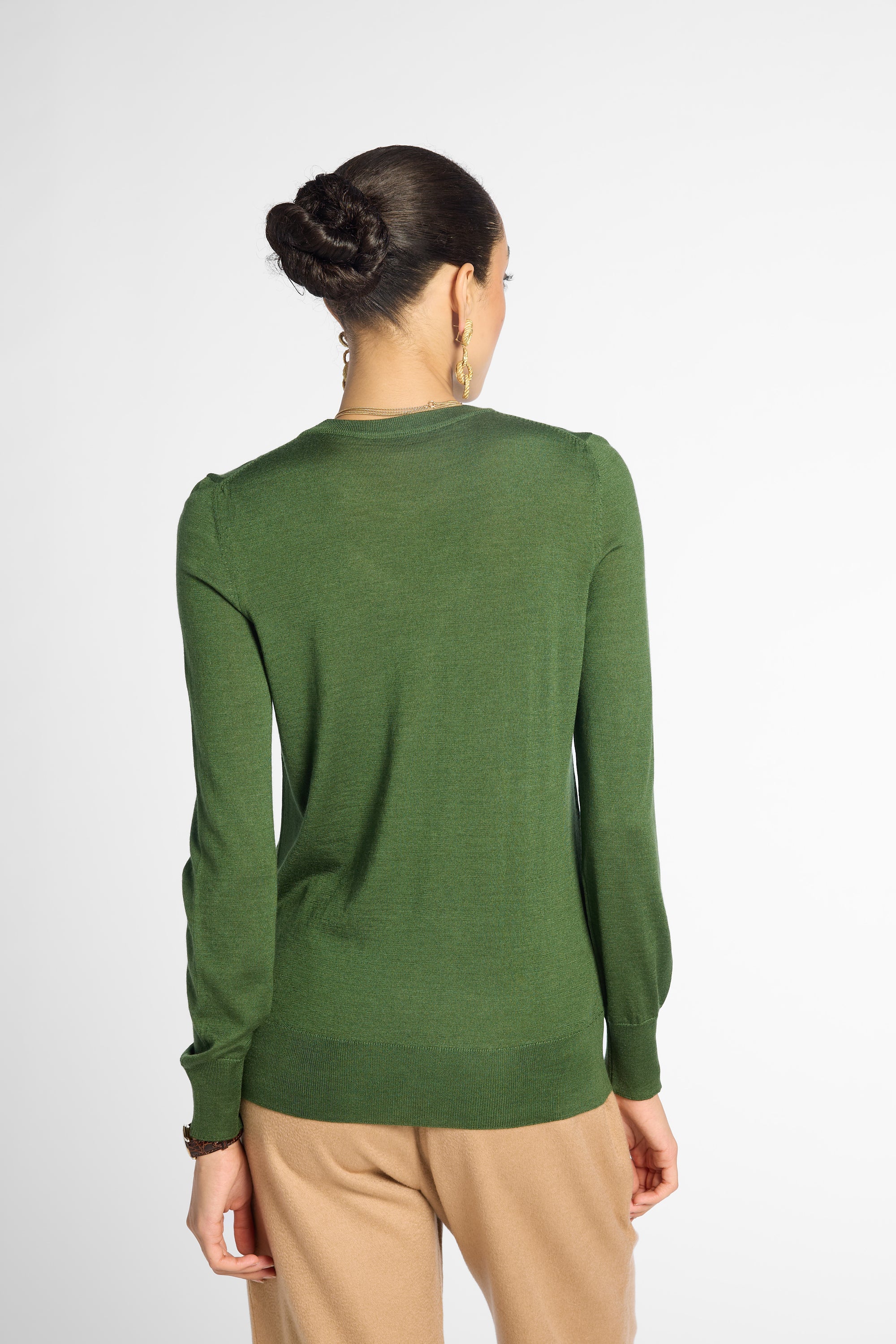 A woman viewed from the back wearing a long-sleeve knit sweater in olive green and tan pants, against a plain light gray studio background.
