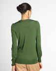 A woman viewed from the back wearing a long-sleeve knit sweater in olive green and tan pants, against a plain light gray studio background.
