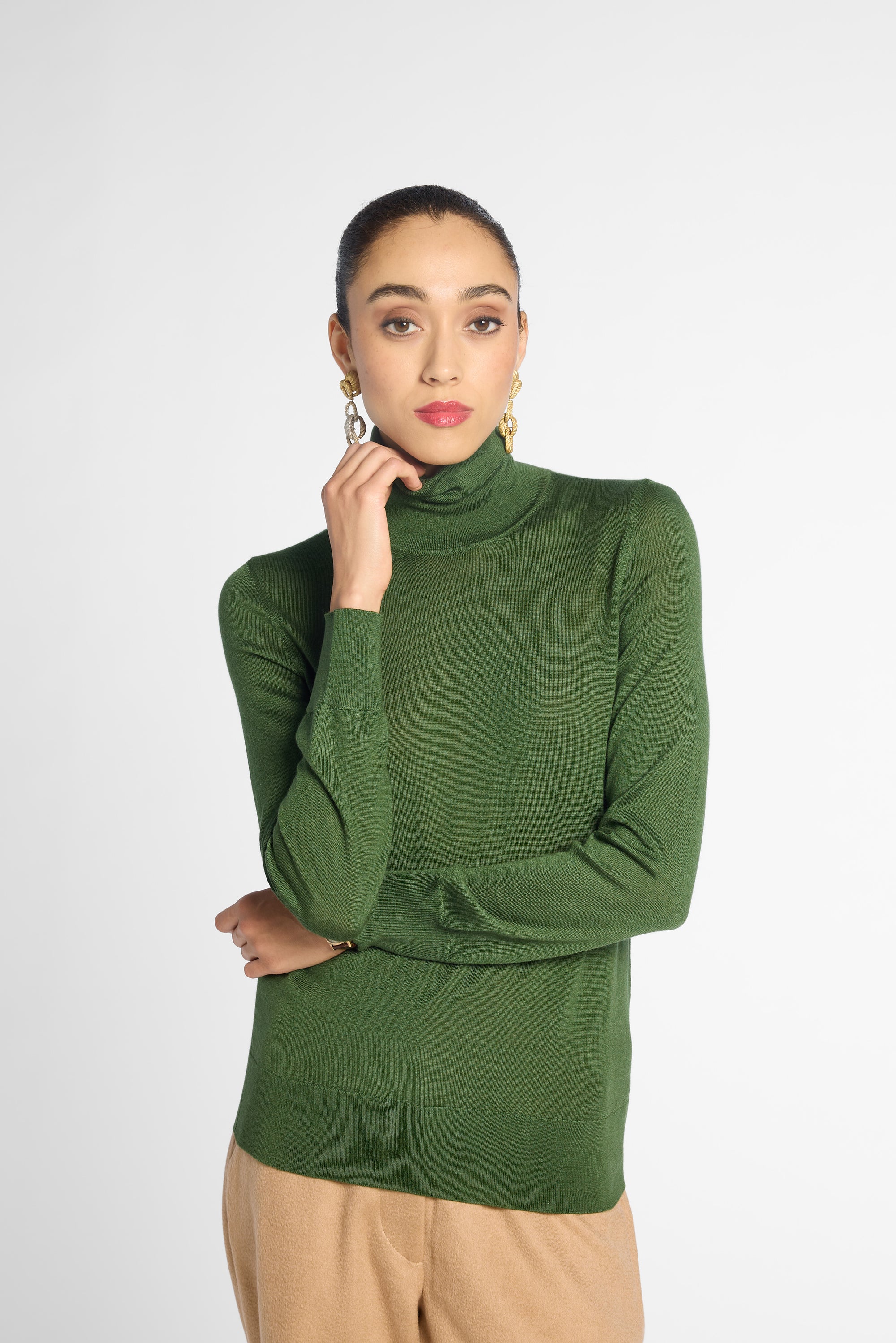A woman wearing a fitted ribbed green turtleneck sweater and beige trousers, standing against a light gray studio background.