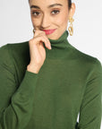 A woman wearing a fitted green turtleneck sweater, styled with gold drop earrings, against a plain light gray studio background.
