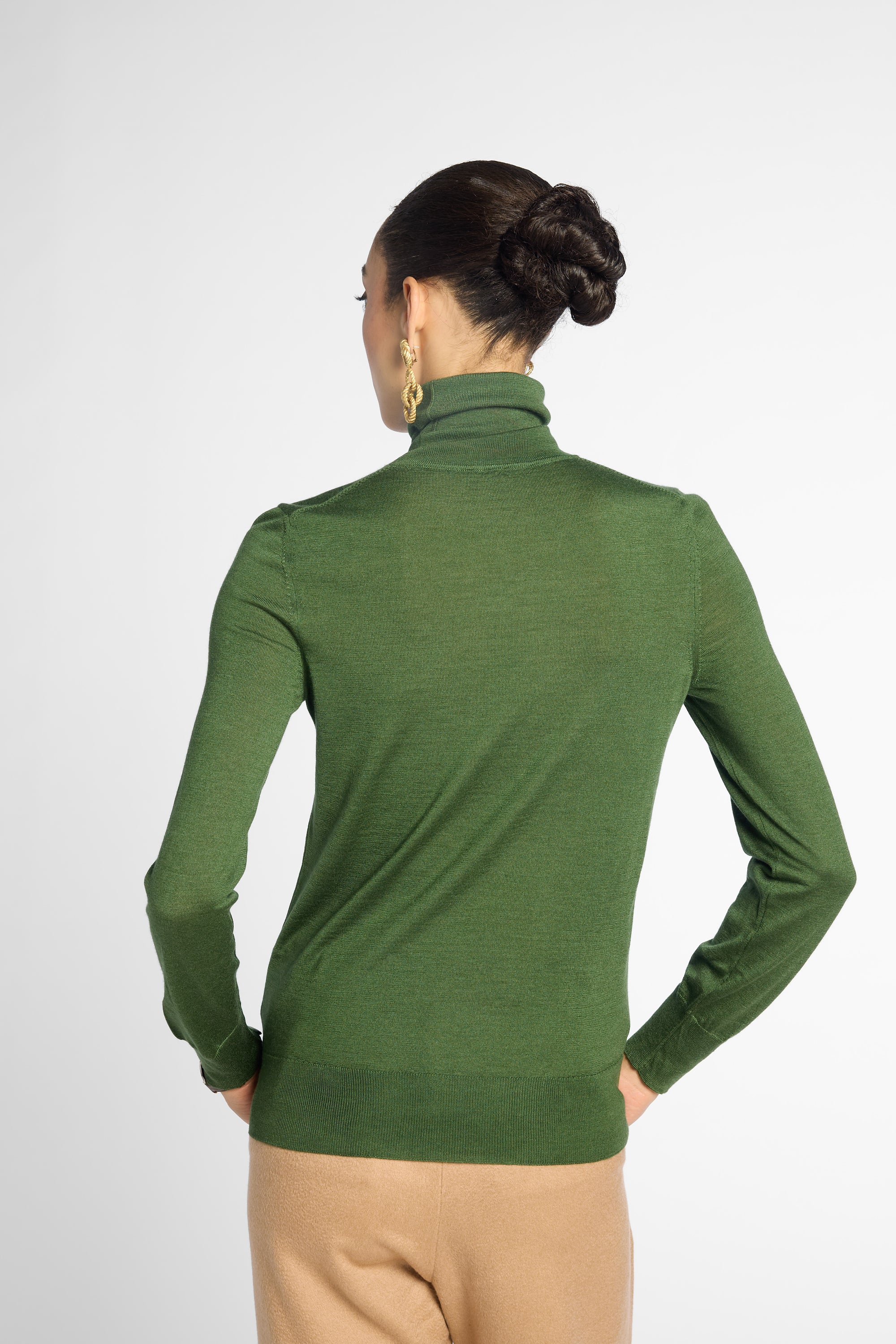 A woman shown from the back wearing a fitted green turtleneck sweater and beige pants against a light gray studio background.