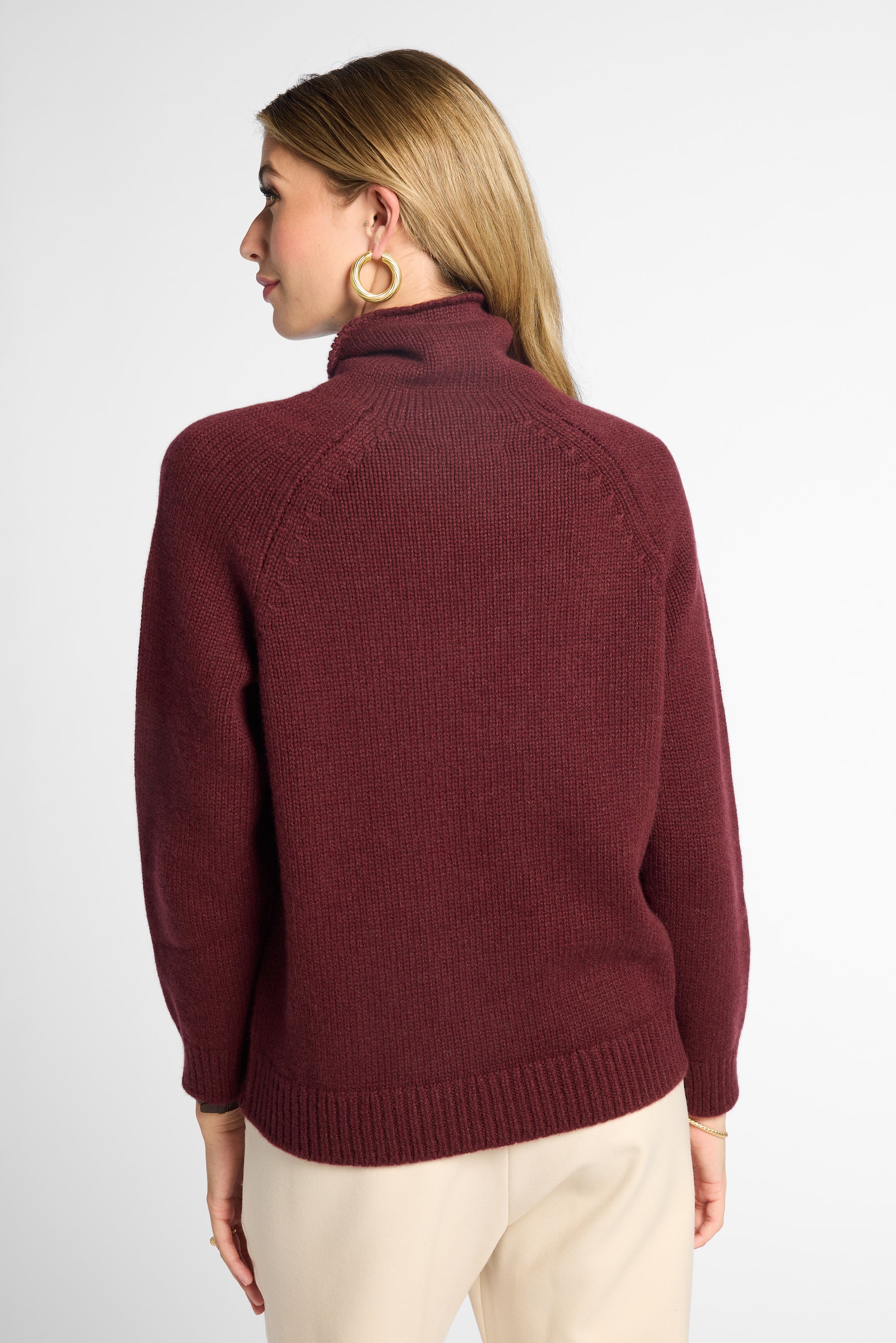 A woman shown from the back wearing a burgundy knit turtleneck sweater and cream pants against a light gray studio background.