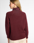 A woman shown from the back wearing a burgundy knit turtleneck sweater and cream pants against a light gray studio background.