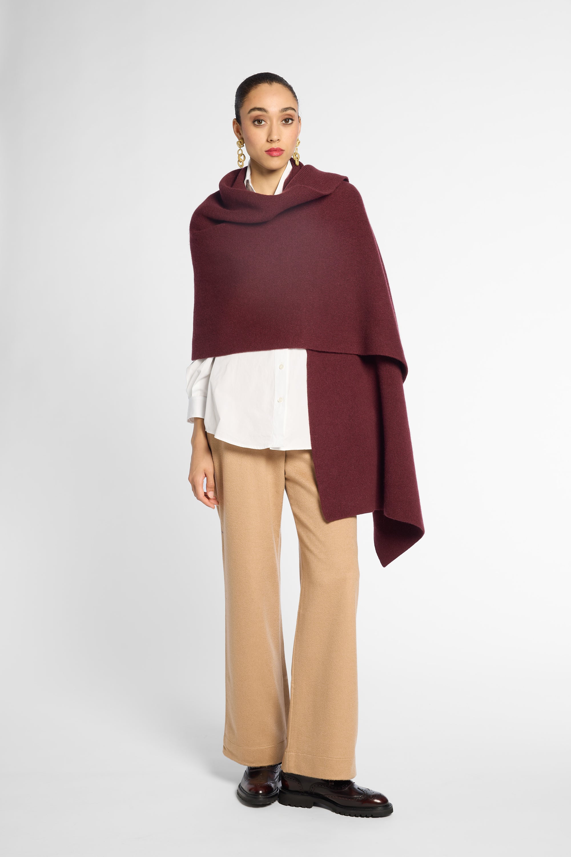 A model stands against a light gray studio background wearing a draped burgundy wrap or poncho over a white collared shirt, beige wide-leg pants, and black shoes.
