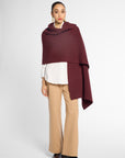 A model stands against a light gray studio background wearing a draped burgundy wrap or poncho over a white collared shirt, beige wide-leg pants, and black shoes.