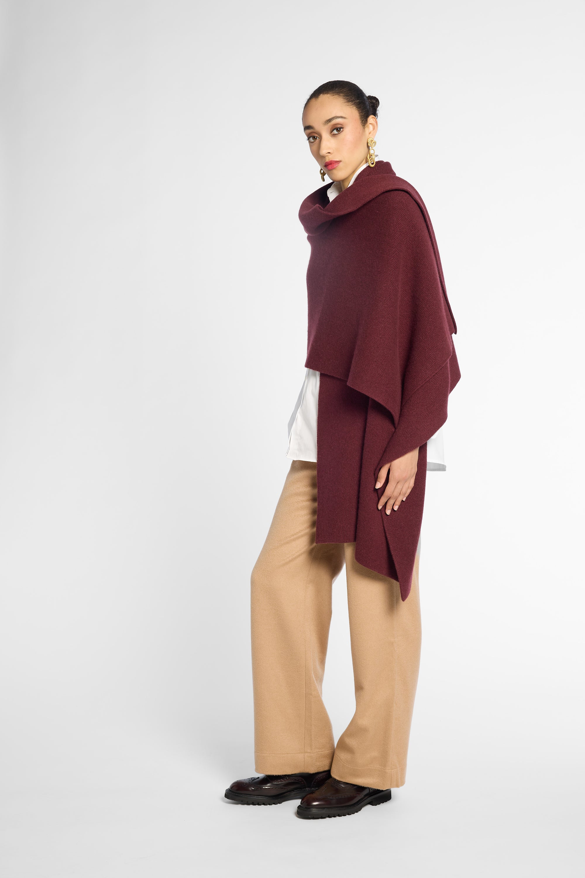 Side view: A woman wearing a chunky knit burgundy shawl draped over her shoulders, styled over a white top, against a plain light gray studio background