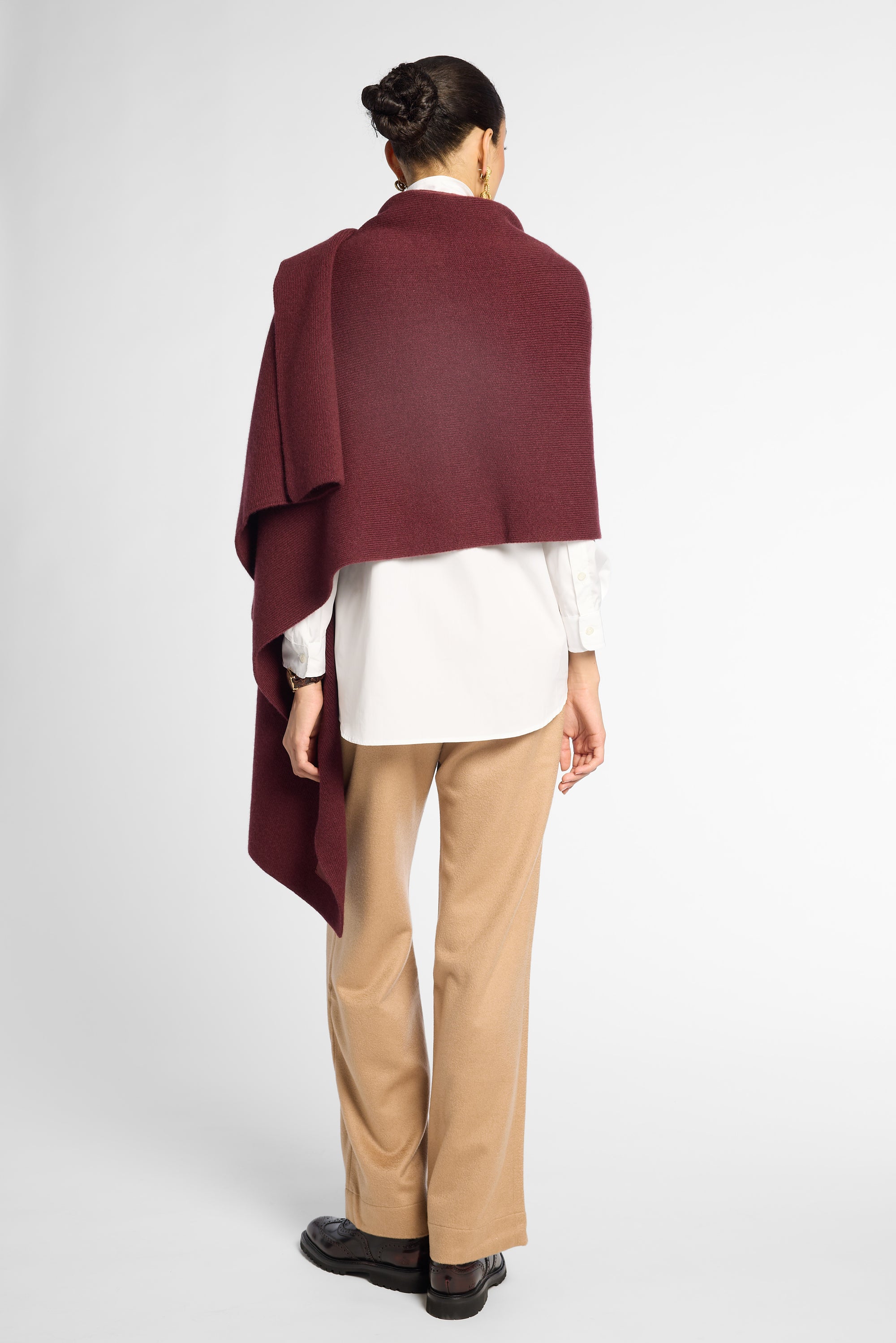 A woman shown from the back wearing a deep burgundy draped wrap or shawl over a white blouse, tan pants, and black shoes, against a light gray studio background.